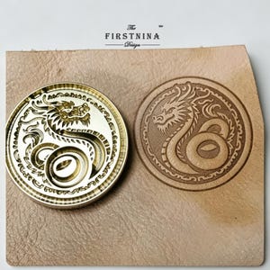 May include: A circular brass metal stamp featuring an engraved dragon design, positioned next to a matching dragon impression on a piece of light tan leather. The text "The FIRSTNINA Design" is printed in black above the stamp and leather.