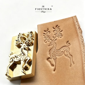 May include: A rectangular brass metal stamp featuring an intricate deer design with ornate antlers, positioned next to a piece of light tan leather showing a clear, debossed impression of the same deer motif. The text THE FIRSTNINA Design is visible.