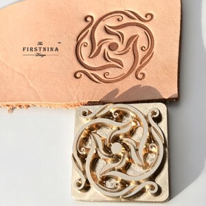 May include: A square metal stamp with a swirling, symmetrical floral pattern and a matching embossed impression on a piece of light tan leather. The text "The FIRSTNINA Design" is printed in black ink on the leather surface near the stamp design.