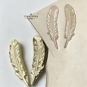 May include: Two gold-colored metal feather stamps and a piece of light beige leather featuring two embossed feather impressions. The text THE FIRSTNINA Design is printed in black ink on the leather surface near the stamped feather designs.