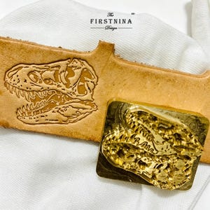 May include: A gold-toned metal stamp featuring a detailed Tyrannosaurus rex skull design, positioned next to a piece of tan leather embossed with the same dinosaur skull image. The text "The FIRSTNINA Design" is printed above the leather piece.