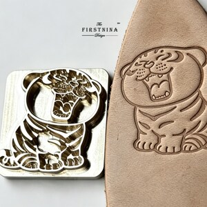 May include: A square brass metal stamp featuring a cartoon tiger design with an open mouth, positioned next to a piece of light tan leather embossed with the same tiger image. The stamp includes the text THE FIRSTNINA DESIGN at the top.