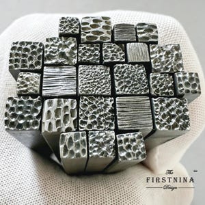 Op de afbeelding: Een verzameling van vijfentwintig vierkante metalen stempelgereedschappen gerangschikt in een raster. Elk gereedschap heeft een uniek getextureerd oppervlak, inclusief diverse gehamerde, gepitte en lineaire patronen. De tekst "The FIRSTNINA Design" staat rechtsonder.