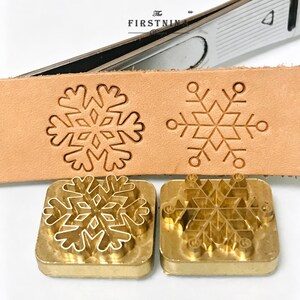 May include: Two square brass metal stamps featuring snowflake designs, positioned below a strip of light tan leather embossed with the same two snowflake patterns. The text "The FIRSTNINA" is visible on a metal tool handle in the background.