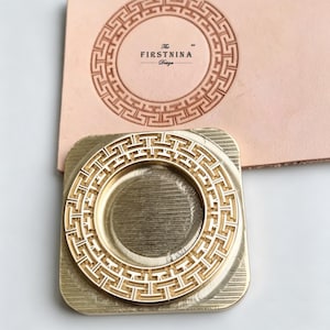 May include: A square brass metal stamp featuring a circular Greek key pattern. The stamp is positioned on a light tan leather surface that displays a clear, embossed impression of the same circular Greek key design and the text The FIRSTNINA Design.