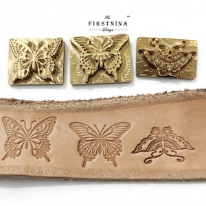 May include: Three rectangular brass metal stamps featuring intricate butterfly designs, positioned above a strip of light tan leather showing the corresponding embossed butterfly impressions. The text "The FIRSTNINA Design" is printed above the stamps.