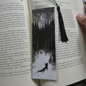 May include: A rectangular bookmark with a black and white illustration of a dark forest, a crescent moon, and two animals. The bookmark is made of a clear material, features a black tassel at the top, and is placed inside an open hardcover book.