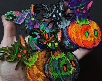 May include: A collection of five metal enamel pins featuring cats wearing pointed hats, each emerging from a pumpkin. The pins display vibrant colors including neon green, orange, purple, and black, with glitter accents and crescent moon details.