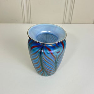 May include: A decorative glass vase with a flared rim and a rounded body. The vase features a blue interior and a vibrant exterior design of swirling red, orange, and teal stripes. The rim is light blue with a gold trim.