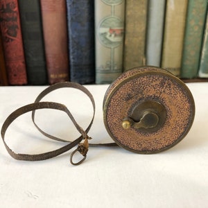 May include: An antique brown and brass measuring tape with a leather tape and a circular case. The case has a textured, aged appearance. The tape is extended, showing its length. The background features a row of vintage books.