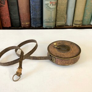 May include: Antique brown and gold tape measure with a leather strap. The tape measure is round with a textured surface and a brass knob. The leather strap is looped and has a metal ring at the end.