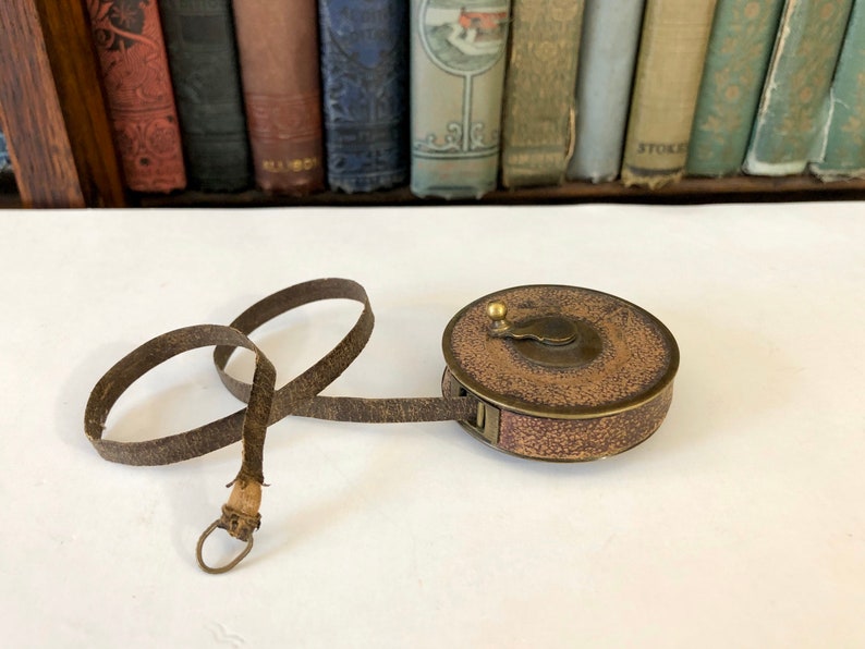 May include: Antique brown and gold tape measure with a leather strap. The tape measure is round with a textured surface and a brass knob. The leather strap is looped and has a metal ring at the end.
