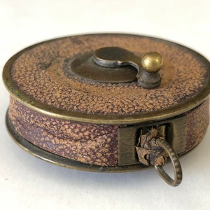 May include: A vintage brown and purple leather tape measure with brass accents. The circular case has a textured surface and a small brass knob on top. A metal ring is attached to the side.