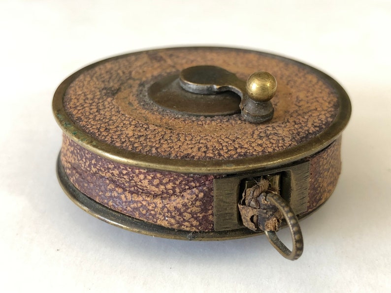 May include: A vintage brown and purple leather tape measure with brass accents. The circular case has a textured surface and a small brass knob on top. A metal ring is attached to the side.