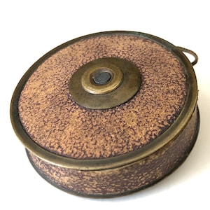 May include: A vintage, round, brown and brass object with a textured surface. The object has a circular design with a central brass piece and a small handle. The brass rim and accents give it an antique appearance.