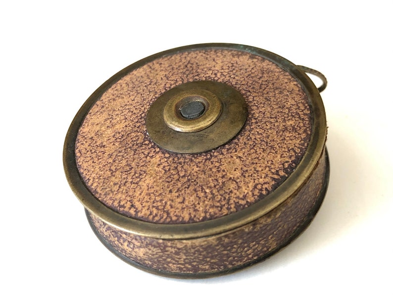 May include: A vintage, round, brown and brass object with a textured surface. The object has a circular design with a central brass piece and a small handle. The brass rim and accents give it an antique appearance.