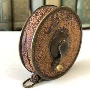 May include: An antique fishing reel with a textured, tan leather exterior and a dark purple interior. The reel has a brass frame and a small, decorative ring attached to the bottom. The reel is a vintage item.