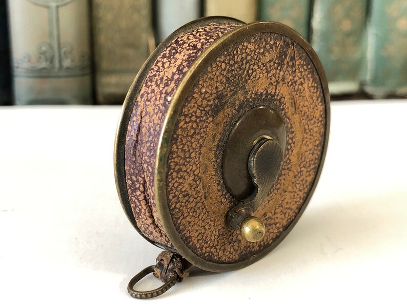 May include: An antique fishing reel with a textured, tan leather exterior and a dark purple interior. The reel has a brass frame and a small, decorative ring attached to the bottom. The reel is a vintage item.