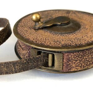 May include: A vintage brown leather measuring tape with a brass handle and trim. The tape is housed in a round case with a textured surface. The leather tape extends from the case, showing signs of wear.