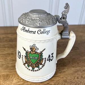 May include: Cream-colored ceramic beer stein with a metal lid and handle. The stein is decorated with the words "Amherst College" and a crest with the letters "ΑΔΦ" and the numbers "19" and "43". The handle features a decorative lion.