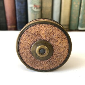 May include: A vintage brass and brown tape measure with a textured face. The tape measure is circular with a central button and a brass rim. The background features a row of antique books.