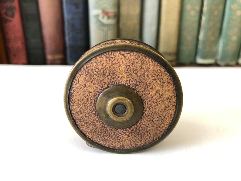 May include: A vintage brass and brown tape measure with a textured face. The tape measure is circular with a central button and a brass rim. The background features a row of antique books.