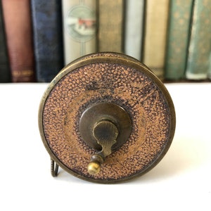 May include: Antique brass and leather tape measure. The tape measure has a circular design with a textured brown leather center and a brass handle. The background features a collection of vintage books.