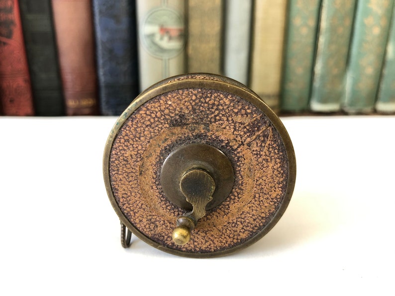 May include: Antique brass and leather tape measure. The tape measure has a circular design with a textured brown leather center and a brass handle. The background features a collection of vintage books.