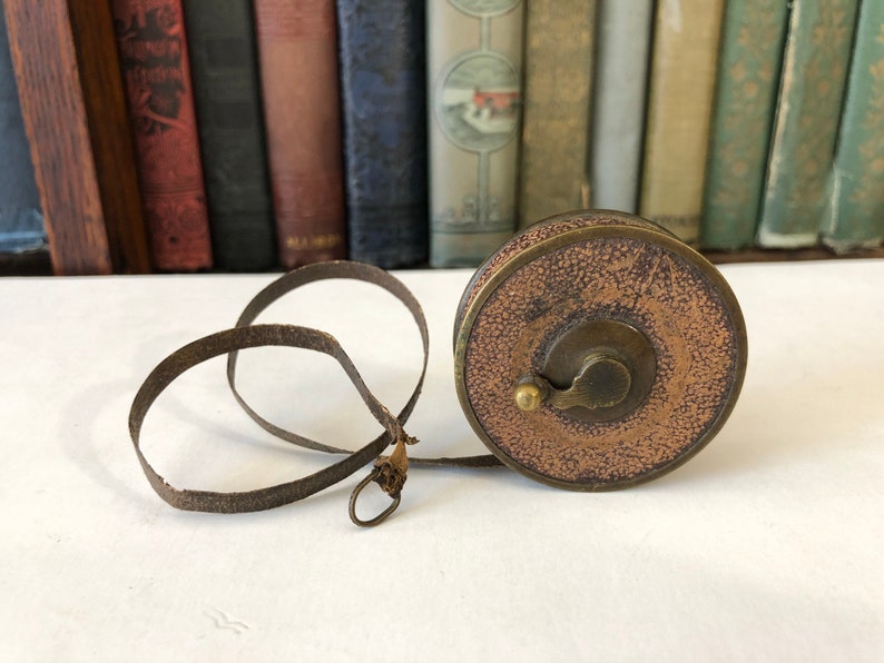 May include: An antique brown and brass measuring tape with a leather tape and a circular case. The case has a textured, aged appearance. The tape is extended, showing its length. The background features a row of vintage books.