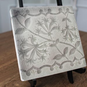 May include: Square, off-white ceramic tile with a gray floral pattern. The tile is supported by a black easel. The floral design includes stylized leaves and flowers, with a decorative border. The tile has a crackled surface.