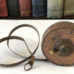 May include: A vintage brown leather tape measure with a brass casing. The tape measure has a textured leather exterior and a metal winding mechanism. The measuring tape itself is a dark brown color and is extended in a loop.