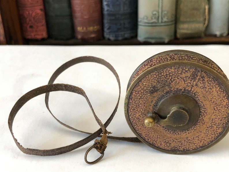 May include: A vintage brown leather tape measure with a brass casing. The tape measure has a textured leather exterior and a metal winding mechanism. The measuring tape itself is a dark brown color and is extended in a loop.