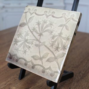 May include: Square ceramic tile with a floral design in shades of gray and white. The tile is displayed on a black easel, showcasing its intricate botanical pattern. The tile's edges have a decorative border.