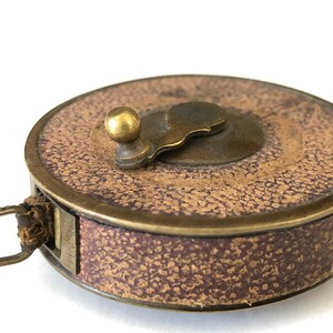 May include: A vintage brown and brass measuring tape with a textured surface. The tape measure has a brass winding handle and a leather loop for carrying. The tape measure is a classic tool.