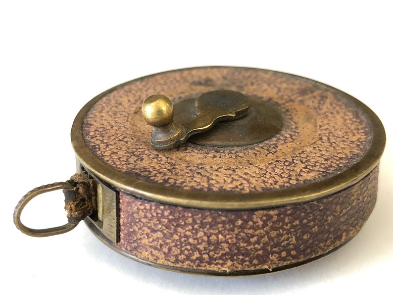 May include: A vintage brown and brass measuring tape with a textured surface. The tape measure has a brass winding handle and a leather loop for carrying. The tape measure is a classic tool.