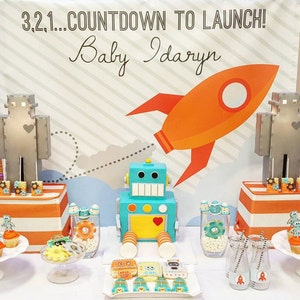 May include: A baby shower display with a robot theme. The backdrop reads "3.2.1... COUNTDOWN TO LAUNCH! Baby Idaryn" with a rocket graphic. A blue robot cake, robot cookies, and other treats are on the table.