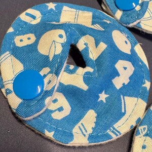 Pu&ograve; includere: Un cerchio di tessuto blu e bianco con un foro al centro. Il tessuto ha un motivo di lettere e forme bianche su uno sfondo blu. C'&egrave; un bottone blu sul lato del cerchio.