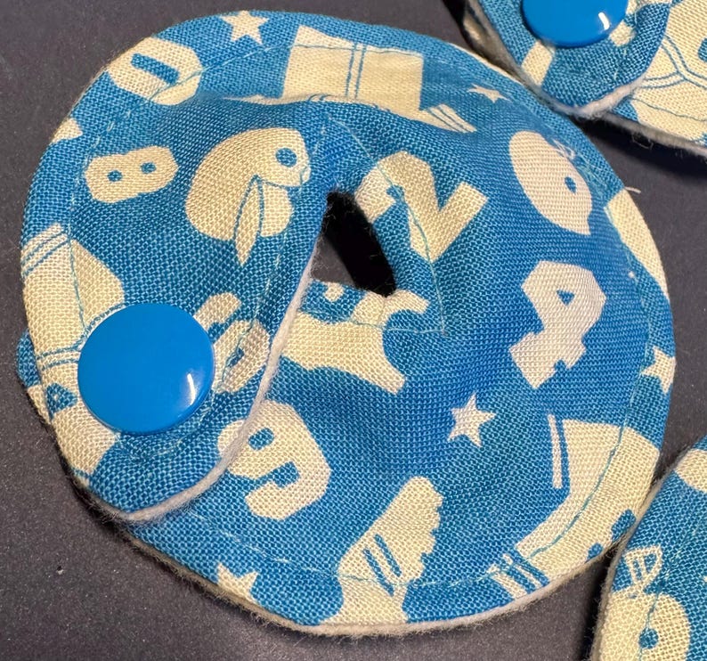 Pu&ograve; includere: Un cerchio di tessuto blu e bianco con un foro al centro. Il tessuto ha un motivo di lettere e forme bianche su uno sfondo blu. C'&egrave; un bottone blu sul lato del cerchio.