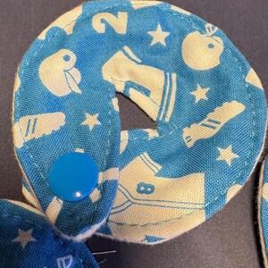 Pu&ograve; includere: Due cerchi di tessuto blu con disegni bianchi, tra cui stelle, un numero 2 e una scarpa. I cerchi sono collegati da un bottone blu.