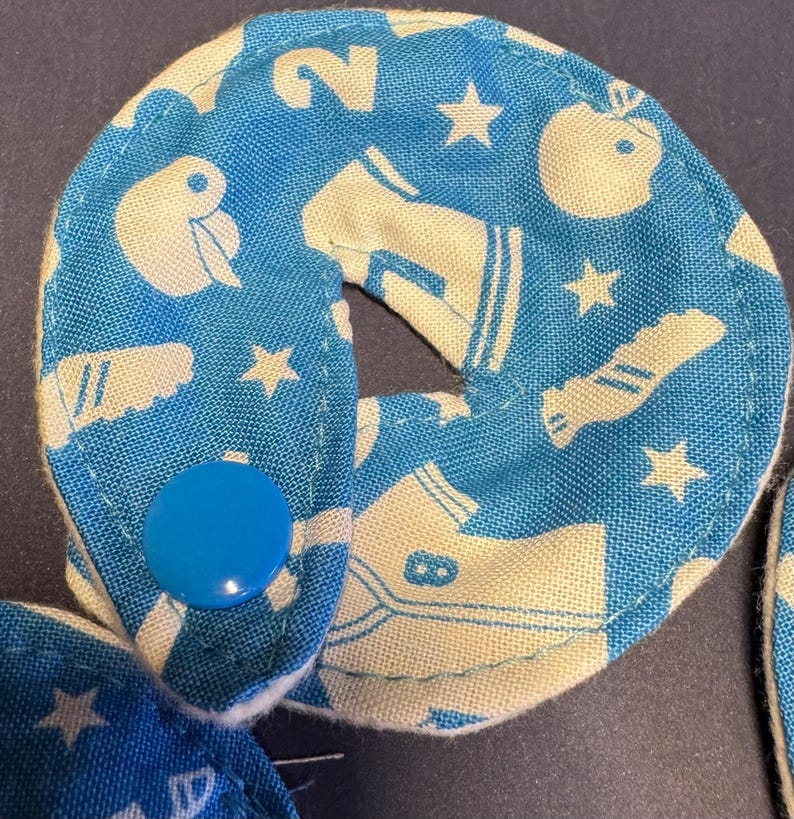 Pu&ograve; includere: Due cerchi di tessuto blu con disegni bianchi, tra cui stelle, un numero 2 e una scarpa. I cerchi sono collegati da un bottone blu.