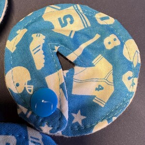 Pu&ograve; includere: Un cerchio di tessuto blu e bianco con un bottone bianco al centro. Il tessuto ha un motivo di immagini legate allo sport, tra cui maglie da baseball, caschi da football americano e scarpe da ginnastica.