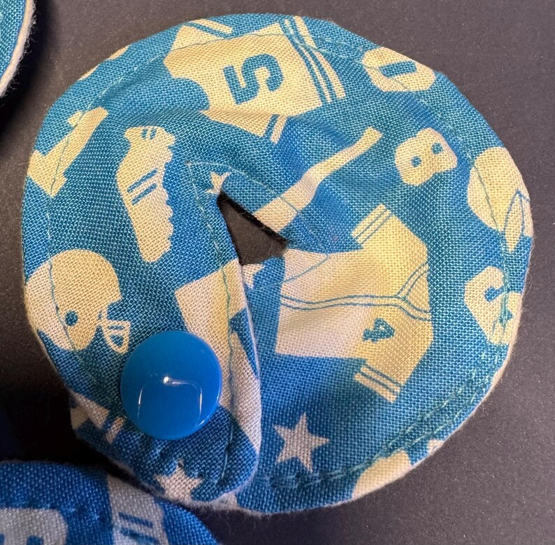 Pu&ograve; includere: Un cerchio di tessuto blu e bianco con un bottone bianco al centro. Il tessuto ha un motivo di immagini legate allo sport, tra cui maglie da baseball, caschi da football americano e scarpe da ginnastica.