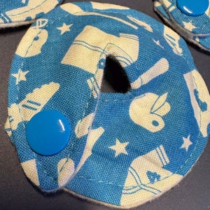 Pu&ograve; includere: Un cuscinetto in tessuto blu con un design bianco di palline da baseball, stelle e scarpe. Il cuscinetto ha un foro al centro e un bottone blu sul lato.