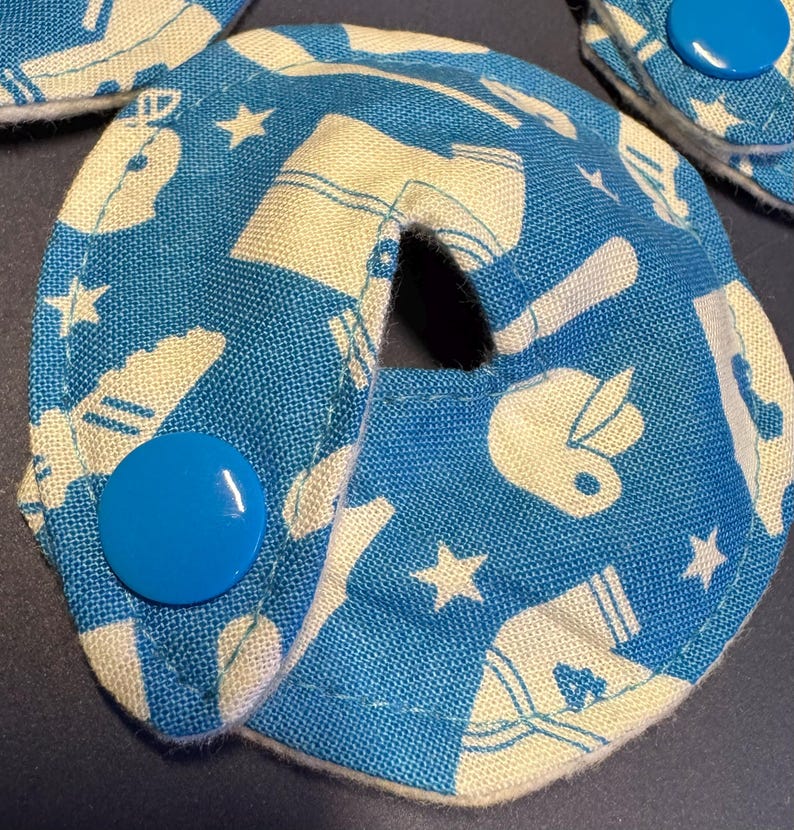 Pu&ograve; includere: Un cuscinetto in tessuto blu con un design bianco di palline da baseball, stelle e scarpe. Il cuscinetto ha un foro al centro e un bottone blu sul lato.