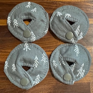 May include: Four gray fabric nursing pads with a white tree pattern and a snap closure. The pads are circular with a hole in the center and are arranged on a wooden surface.
