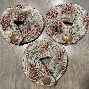 May include: Three brown and green fabric coasters with a pine cone pattern and a brown button in the center of each coaster.
