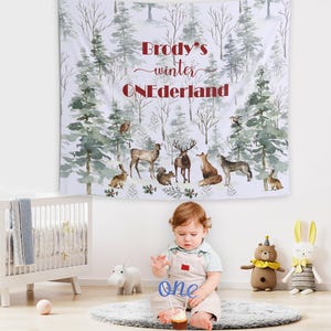 May include: A white tapestry with a winter forest scene and the text "Brody's winter ONEDerland." The tapestry features illustrations of trees and woodland animals. A baby sits on a rug holding a "one" cake topper.