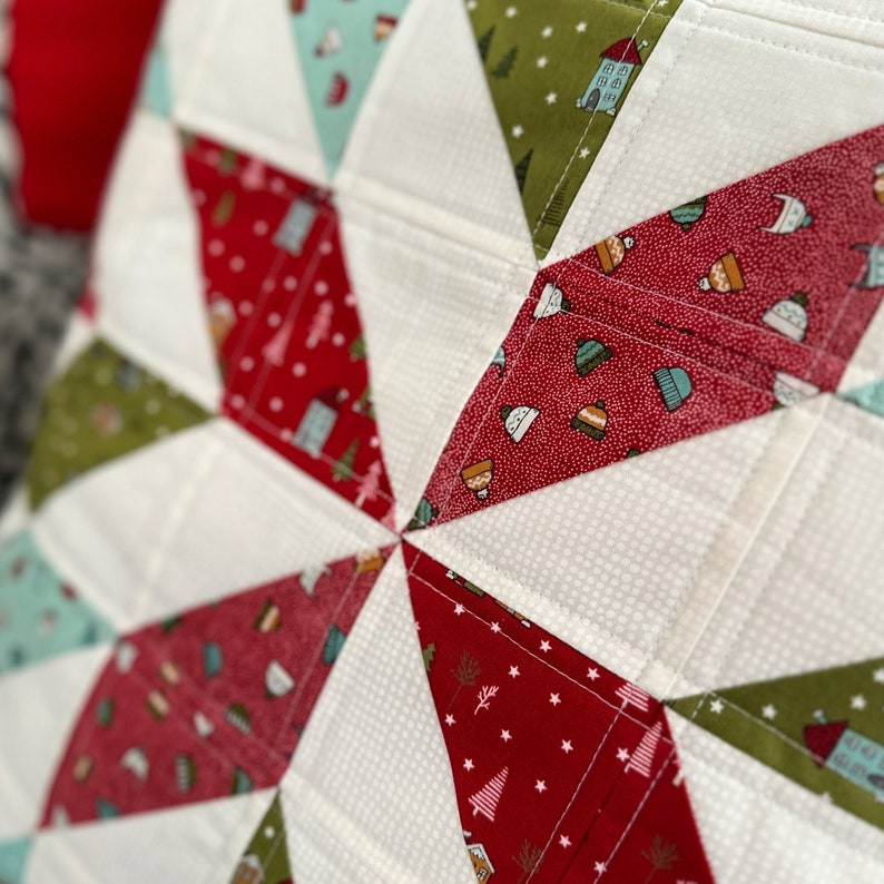 May include: Close-up of a quilted textile with a starburst design. The quilt features red, green, and white triangular sections. The red sections have small house and tree patterns. The green sections have a similar pattern. The white sections have a dotted pattern.