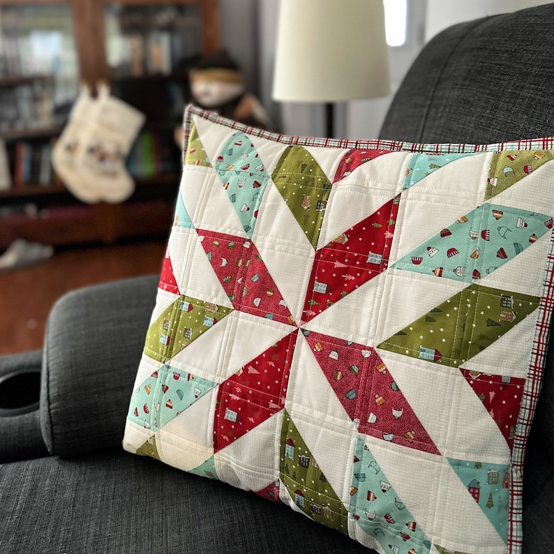 May include: A square decorative pillow with a starburst pattern. The starburst design features red, green, and light blue triangles with small house and tree motifs, set against a white background. The pillow has a red and white plaid border.