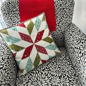 May include: A decorative pillow with a starburst pattern in red, green, and blue geometric shapes. The pillow is on a chair with a black and white floral pattern, and a red throw is draped over the back.